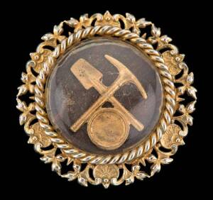 A collection of three Miner's brooches, one with crossed shovels, nuggets and a bucket, stamped 9ct; a circular brooch with crossed pick and shovel, yellow and rose gold; to be sold with a pressed metal costume brooch.