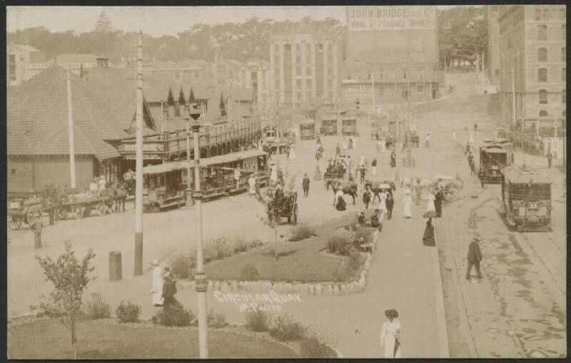 NSW: Sydney: ‘Circular Quay’ real photo postcard showing the Ferry Terminal and Tram Terminus, pedestrians and horse & cart traffic and advertising hoardings for ‘John Bridge and Co’ & ‘Wolfe's Schnapps’ in background, unused, fine condition.