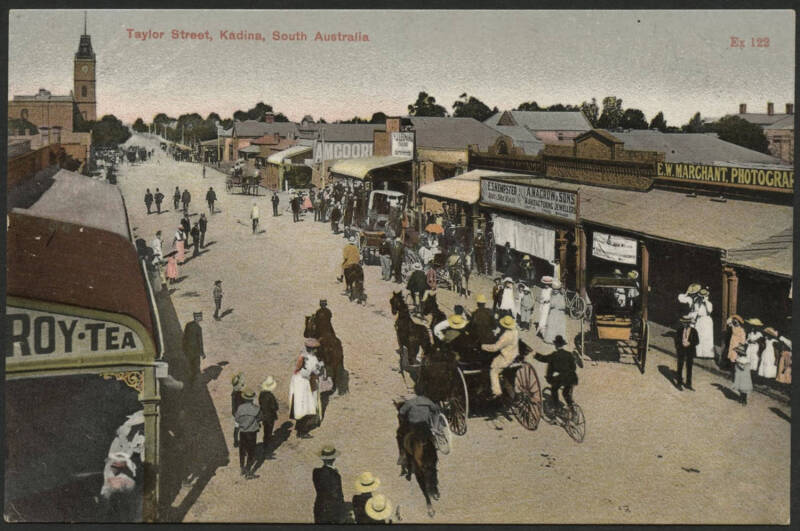 Sth Aust: Kadina: ‘Taylor Street, Kadina’ postcard with storefronts including EW Marchant Photographers, Macrow & Sons Jewellers, ES Kempster Boot & Shoe Maker, also advertising hoardings, horse-drawn carriages, bicycles & pedestrians, unused, fine condit