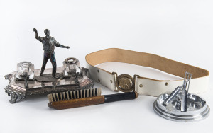 A white metal ashtray featuring crossed bats leaning against a wicket; and, a silver-plated desk inkwell set featuring a bowler, circa 1910 (with minor faults).  Also, a brass belt buckle depicting 3 bats leaned against a tree stump and a clothes brush in