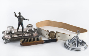 A white metal ashtray featuring crossed bats leaning against a wicket; and, a silver-plated desk inkwell set featuring a bowler, circa 1910 (with minor faults). Also, a brass belt buckle depicting 3 bats leaned against a tree stump and a clothes brush in the shape of a cricket bat marked "Souvenir of Australia". An interesting little group. (4 items).