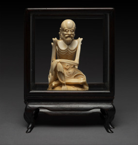 A Chinese carved ivory seated skeletal figure in timber and glass case, 19th/20th century, seal mark to base, ​the figure 12.5cm high