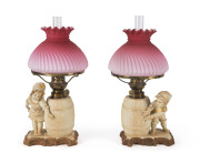 A pair of German figural bisque porcelain kerosene lamps with ruby glass shades, 19th century, 34cm high