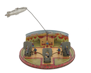 "Coast Defence" wind-up tinplate toy by Louis Marx & Co (USA), the wind-up mechanism concealed within a defence building which has a metal rod extending from the roof allowing a detachable zeppelin to circle above, in the foreground are three manned gun stations which move back and forth when the mechanism is activated. Circa.1930s; toy size 22½cm (9") diameter, height 17cm (7") when dirigible is attached, (damaged) box size 23½cm (9.5") x23cm (9.5").