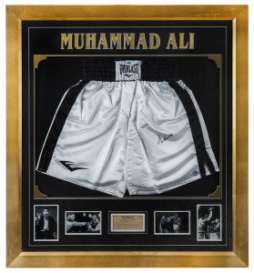 MUHAMMAD ALI: deluxe timber framed display comprising pair of signed 'Everlast" boxing shorts with four inset black & white iconic images of Ali together with a plaque providing his fight chronology, Ceritificate of Authenticity on reverse together with a photograph of Ali signing one of the 125 pairs of boxing shorts he signed on 12th & 13th June, 1998 in New York City; overall 103x113cm.