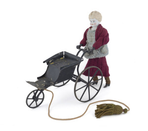 Antique pull-along doll pushing a three-wheel carriage; appears to be based on the Godwin clockwork walking doll by Stevens & Brown of Connecticut (circa 1870s) but does not have the mechanism under the seat of the carriage. The doll, with ceramic head and torso, has metal legs and feet in her original shoes and they are fully functional providing a quite realiztic walking movement; the clothing appears to be a combination of original garments and later additions. Height: 28.5cm (11.25").