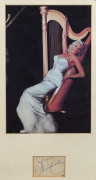 JAYNE MANSFIELD: mounted display featuring a colour photograph of platinum blonde Mansfield in evening dress leaning against a harp, signature on a plain piece beneath; framed & glazed, overall 47x30cm.