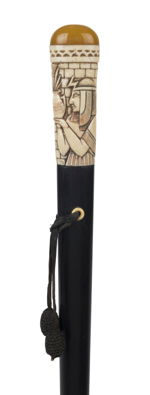 An antique walking stick, carved ivory handle with Egyptian motif topped with polished amber, tapering ovoid shaft in ebony with brass ferrule, circa 1900, 96.5cm high