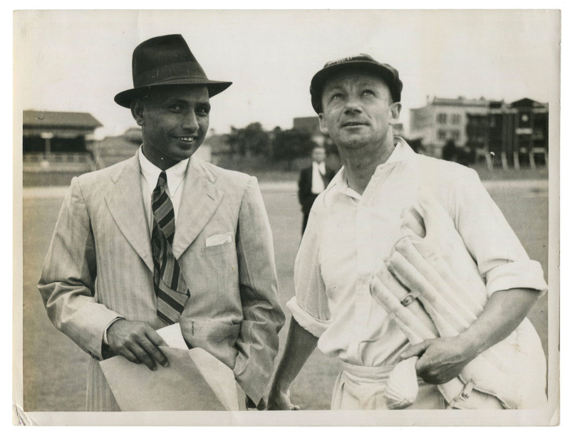 DON BRADMAN - AUSTRALIA vs INDIA, 1st TEST, BRISBANE, Nov.28 - Dec.2, 1947: original press photograph (The Courier Mail, Brisbane), showing Bradman on the ground in his cricket gear looking skyward alongside a besuited Indian Captain Lala Armanath; 21.5 x