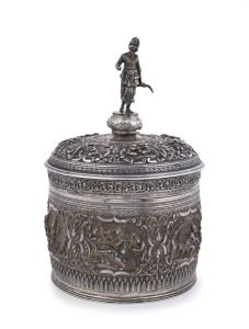 A stunning antique Burmese silver ceremonial betel box. Decorated with six deep repoussé cartouches showing Burmese mythological scenes with lotus and scroll frieze. The lid also beautifully worked in repoussé with seven further figural vignettes and a spectacular cast finial figure of HANUMAN holding his tail. Lower Burma, circa 1890. 24cm high, 15.5cm diameter, 958 grams.