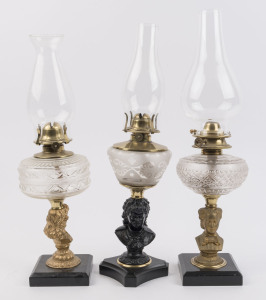 Three kerosene lamps with cast bust stands on ebonised cast metal bases, pressed glass fonts, one with frosted decoration, all with single brass burners and glass chimneys, late 19th century, (3 items), the largest 56cm high