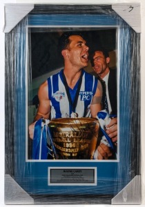 NORTH MELBOURNE - WAYNE CAREY: mounted large colour photograph (45x30cm) of Carey holding the 1996 Premiership Cup, SIGNED BY CAREY, commemorative plaque beneath highlighting his career achievements, with CofA; framed and glazed, overall 67x45cm.
