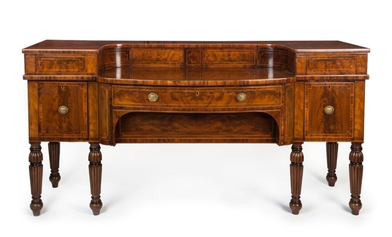 An impressive antique English mahogany bow front sideboard, beautifully crafted with handsome cuts of flame mahogany, string inlay, cross banding, six turned and reeded legs and original brass drop handles, early 19th century. One of the finest examples s