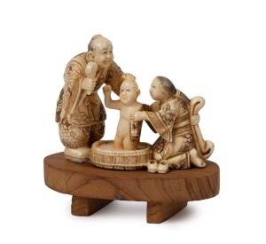 KYOTO SCHOOL finely carved ivory netsuke of two parents washing a child, Meiji period, 19th century, mounted on a timber stand, 3.5cm high, 4.5cm wide, 5cm high with stand