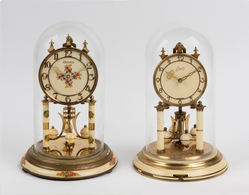 Two German 400 day anniversary clocks in glass and perspex domes with cream enamel decoration, 20th century, 29cm and ​​​​​​​30cm high each overall