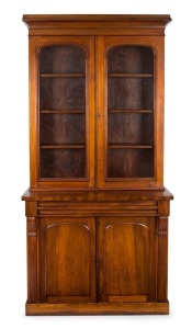 An Australian cedar bookcase with a cavetto pediment, two arch-top glazed doors, conforming lower panelled doors, an ogee-moulded drawer and carved corbels, 19th century, 235cm high, 114cm wide, 44cm deep