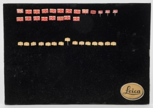 LEITZ: Thirty-six Leica-branded metal pins, twenty-one in red and fifteen in gold, presented on a black Leica-branded pinboard. (37 items)