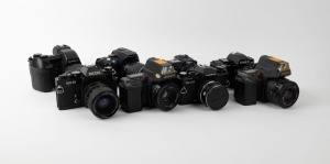 VARIOUS MANUFACTURERS: Eight black SLR cameras - one Canon PowerShot Pro1 with lens hood and lens cap, one Konica Minolta Dynax 5D with lens hood, one Minolta Maxxum 7000 AF with front lens cap, one Olympus Camedia E-10, one Ricoh KR-10, one Voigtländer VSL 3-E with front lens cap, two Yashica Kyocera 230-AF models with differing lenses. (8 cameras)