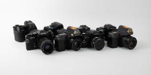 VARIOUS MANUFACTURERS: Eight black SLR cameras - one Canon PowerShot Pro1 with lens hood and lens cap, one Konica Minolta Dynax 5D with lens hood, one Minolta Maxxum 7000 AF with front lens cap, one Olympus Camedia E-10, one Ricoh KR-10, one Voigtländer V