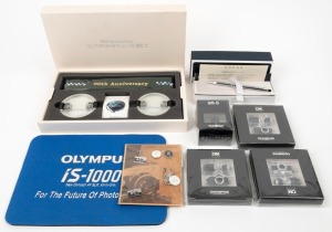 OLYMPUS: Eight promotional items - one 90th Anniversary Olympus Space Project Commemorative Kit comprising two replicas of the Olympus E-3 lens caps designed for use on the International Space Station, with camera strap and information sheet in maker's box, together with four miniatures sealed in retail packaging (two black OM-1, one grey OM-1, and one grey OM-D), one Classic Pen F sealed set of five pins, one metal Cross pen with 'Olympus Pen' imprint, and one iS-1000 mouse pad. (8 items)