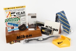 NIPPON KOGAKU: Thirteen Nikon-branded items - one wooden 25x17.5x3.5cm lens stand, one plastic 13x11x3.5cm lens stand, one 11.5x8x10cm Nikon sign, one 6x4x11cm three-dimensional Nikon D300 glass etching, three pin badges, one necktie, one black plastic wristwatch in transparent plastic case, one 1998 50th-anniversary camera strap, and other items. (13 items)