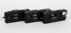 OLYMPUS: Three black-body compact cameras with attached wrist straps - one 1979 Olympus XA [#4465952] with F. Zuiko 35mm f2.8 lens and A16 flash unit, one 1979 Olympus XA-1 [#4802067] with D. Zuiko 35mm f4 lens and A11 flash unit, and one 1982 Olympus XA-