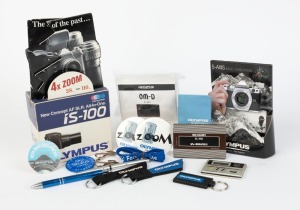 OLYMPUS: Nineteen Olympus-branded items, including one Casio calculator, one Sharp calculator, three keyrings, three metal pens, two tie clips, one plastic OM-D miniature in retail packaging, four badges, two point-of-sale displays, and other items. (19 i