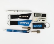 OLYMPUS: Nineteen Olympus-branded items, including one Casio calculator, one Sharp calculator, three keyrings, three metal pens, two tie clips, one plastic OM-D miniature in retail packaging, four badges, two point-of-sale displays, and other items. (19 i - 2