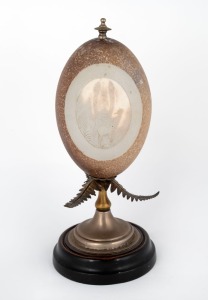 An antique emu egg ornament with carved lyrebird cartouche on silver plated fern mounts on turned ebonized timber base, circa 1900, 24cm high