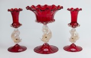 A three piece Murano ruby glass mantle garniture set with dolphin bases, circa 1960s, bearing original foil labels, ​​​​​​​the largest 24cm high.