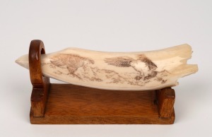 A scrimshaw hippopotamus tooth ornament with engraved eagle decoration, signed lower right with monogram (illegible), mounted on timber stand, 20th century, 16.5cm long