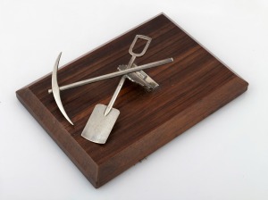 An Australian silver document clip in the form of a crossed pick and shovel, mounted on a timber base, 20th century, ​​​​​​​21 x 15cm overall.
