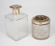 An Art Deco sterling silver lidded crystal bottle of square form, together with a cylindrical antique hobnail cut crystal jar with floral decorated sterling silver lid, 19th/20th century, (2 items), ​​​​​​​14cm and 9cm high, the lids 56 grams total.