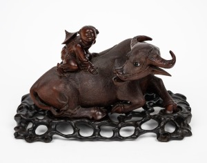 An antique Chinese carved timber figural group of a rider and buffalo, adorned with glass eyes and bone teeth, resting on original carved timber stand, 19th century, 13cm high, 26cm wide overall.