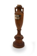 A replica Ashes urn in timber, on matching timber plinth, engraved at base "Made By Alan Kelly Mornington Vic(toria)", with details of the triumphant 1989 Australian team affixed to the base; height 26cm.