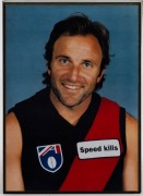 Various framed items including the Herald Sun banner "ABLETT BOWS OUT", a full-page poster of Polly Farmer, an "ABLETT 1000 ADIDAS" poster, an Essendon 1997 team photo, and a photo of Tim Watson in his Essendon jumper; the largest 72 x 52.5cm. (5 items) - 4