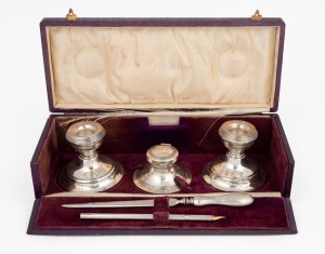 A boxed sterling silver desk set, comprising of an ink pot, candlesticks, pen and burnishing tool, housed in a purple leather silk and velvet lined box, early 20th century, the box 27cm wide.
