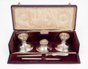 A boxed sterling silver desk set, comprising of an ink pot, candlesticks, pen and burnishing tool, housed in a purple leather silk and velvet lined box, early 20th century, the box 27cm wide.